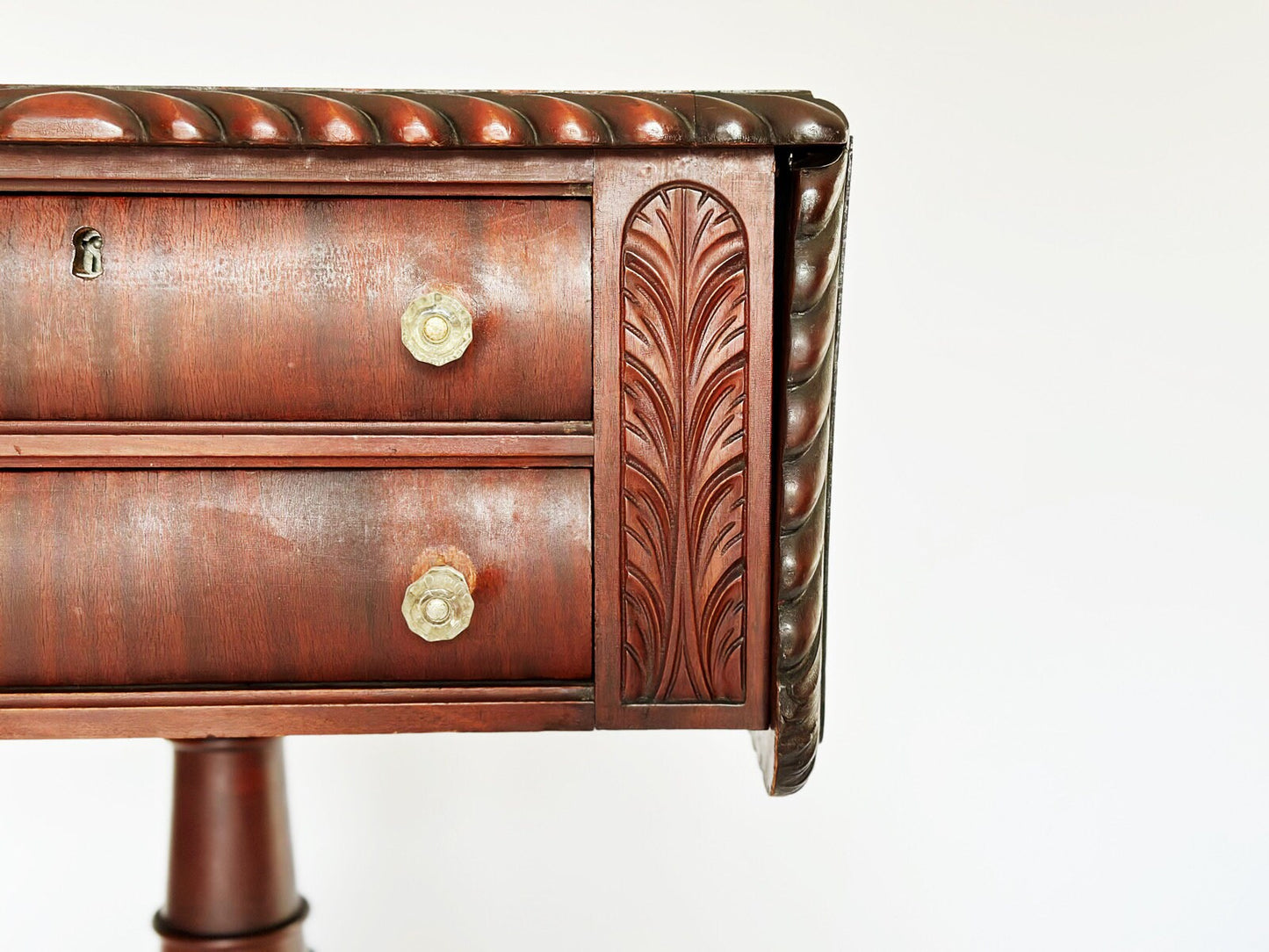 This mahogany antique Empire sewing stand end table has 2 stacked drawers with glass knobs. On either side of the drawers is a vertical carving of a palm leaf frond.