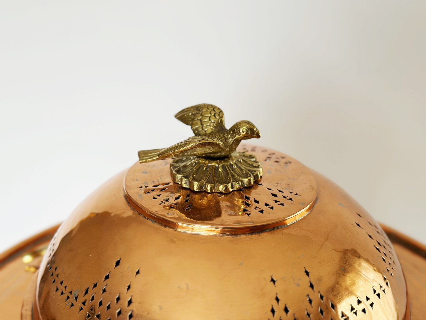 The top vented dome of the vintage Turkish copper brazier fire pit features a decorative brass bird handle to lift off the top. The vented holes of the dome cover form a mix of horizontal and vertical line patterns around the dome.